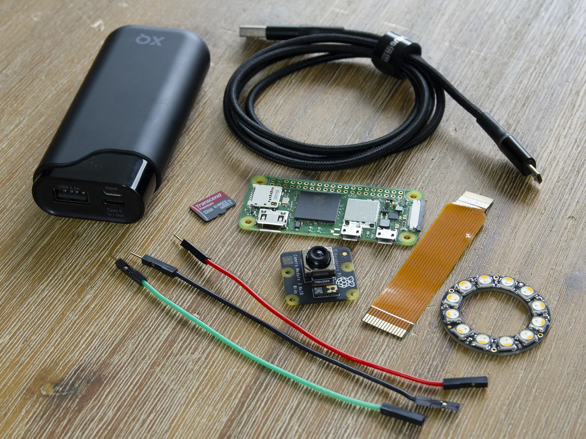 The parts laid out on a workbench: USB power bank, cable, microSD card, Raspberry Pi Zero 2 W, Camera Module 3 Wide, ribbon cable, LED ring and jumper wires