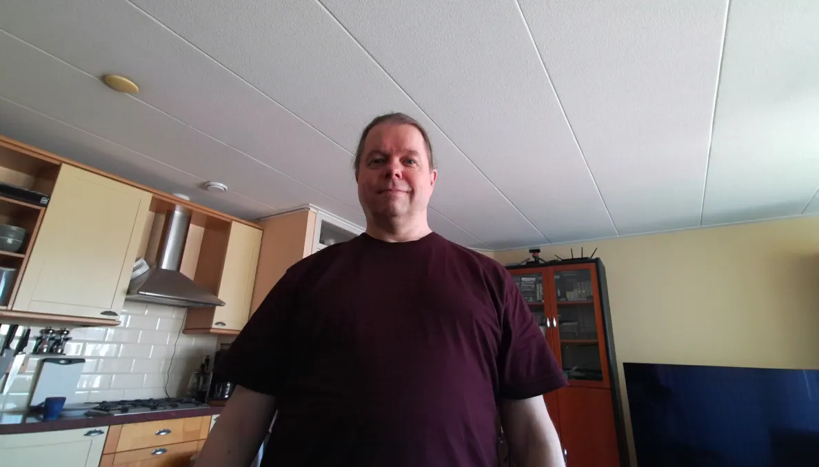 The colour frame from a depth sensor: a man in a dark red t-shirt standing in a kitchen, with cabinets and a cooker hood behind him to the left and a tall glass-doored cabinet and a television to the right